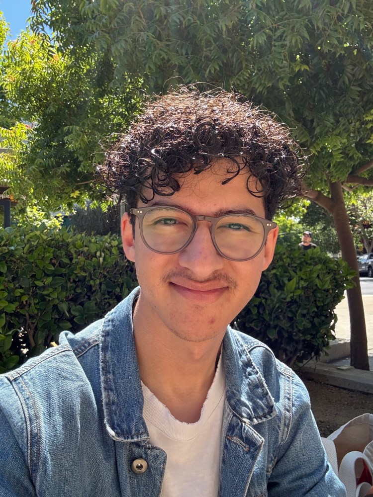 Cropped view of Gabriel Duenas' face and upper torso with round glasses wearing a jean jacket. He is sitting in front of an outdoor garden with shrubs and trees.